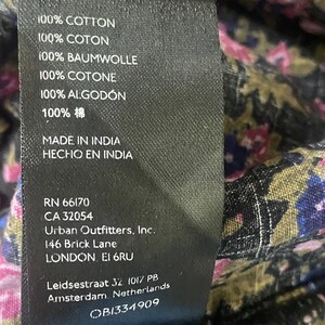 May include: A close-up of a black tag with white text, listing fabric composition as 100% cotton in multiple languages. The tag also includes the brand name, address, and other details. The background shows a floral patterned fabric.