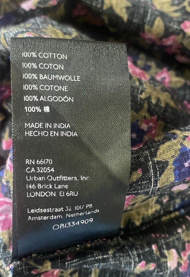 May include: A close-up of a black tag with white text, listing fabric composition as 100% cotton in multiple languages. The tag also includes the brand name, address, and other details. The background shows a floral patterned fabric.