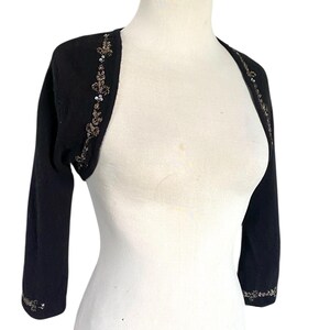 May include: Black bolero sweater with three-quarter sleeves. The sweater features intricate gold and silver sequin and bead embellishments along the edges and upper sleeves. The bolero is displayed on a white mannequin.