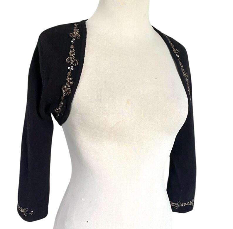 May include: Black bolero sweater with three-quarter sleeves. The sweater features intricate gold and silver sequin and bead embellishments along the edges and upper sleeves. The bolero is displayed on a white mannequin.