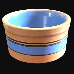 May include: A small, round ceramic bowl with a light brown exterior and a blue interior. The bowl features a blue band with two black stripes. The bowl is likely used for serving dips, sauces, or small portions of food.
