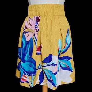 Op de afbeelding: Mosterdgele rok met een bloemenpatroon in blauw, wit en roze. De rok heeft een hoge taille en een textuurstof. Het bloemendesign is een gedurfde, artistieke print. De rok wordt op een mannequin gepresenteerd.