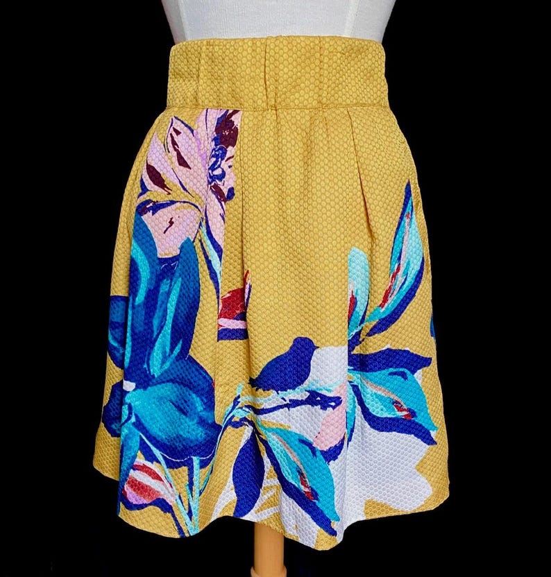 Op de afbeelding: Mosterdgele rok met een bloemenpatroon in blauw, wit en roze. De rok heeft een hoge taille en een textuurstof. Het bloemendesign is een gedurfde, artistieke print. De rok wordt op een mannequin gepresenteerd.