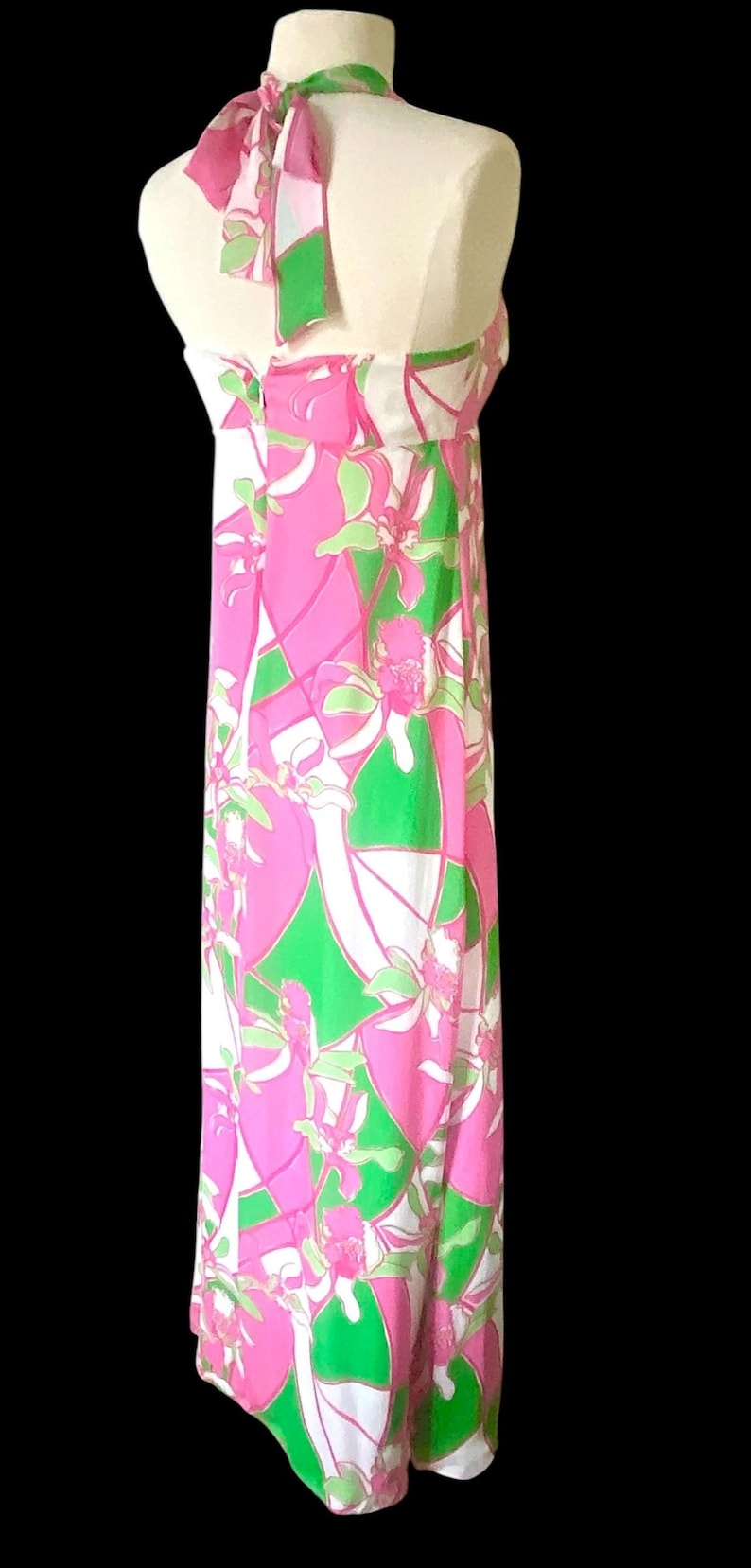 Op de afbeelding: Lange mouwloze jurk met een levendig bloemenpatroon in roze, groen en wit. De jurk heeft een halterhalslijn en een vloeiend silhouet. De stof lijkt lichtgewicht, geschikt voor warm weer.
