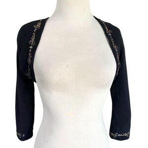 May include: Black bolero sweater with three-quarter sleeves. The edges are adorned with a delicate floral pattern in gold and silver sequins. The sweater is displayed on a white mannequin, showcasing its design and fit.