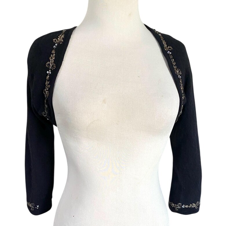 May include: Black bolero sweater with three-quarter sleeves. The edges are adorned with a delicate floral pattern in gold and silver sequins. The sweater is displayed on a white mannequin, showcasing its design and fit.