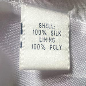 Op de afbeelding: Een wit stoffen label met zwarte tekst: "SHELL: 100% SILK LINING 100% POLY". Het rechthoekige label is in een kledingstuk genaaid, tegen een glanzende, lichtgekleurde stoffen achtergrond.