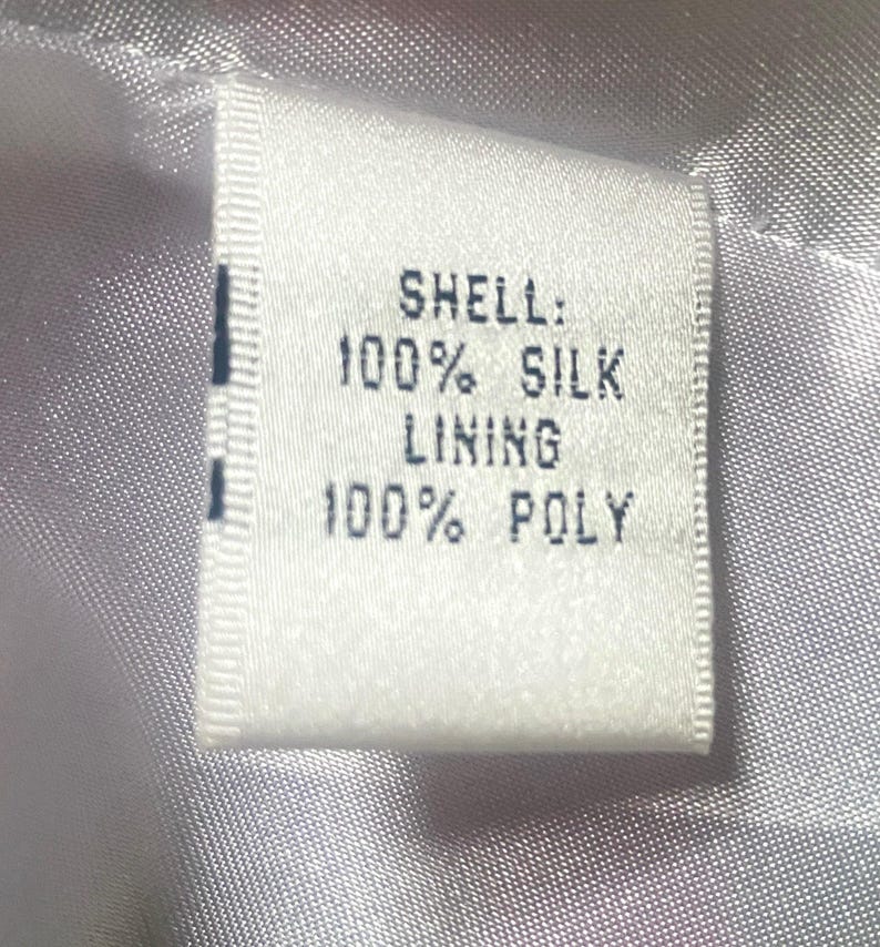 Op de afbeelding: Een wit stoffen label met zwarte tekst: "SHELL: 100% SILK LINING 100% POLY". Het rechthoekige label is in een kledingstuk genaaid, tegen een glanzende, lichtgekleurde stoffen achtergrond.