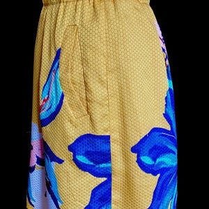 Op de afbeelding: Een gele rok met een bloemenpatroon. De rok heeft grote blauwe en roze bloemen op een gele, getextureerde achtergrond. De rok heeft een getailleerde tailleband en een zak.
