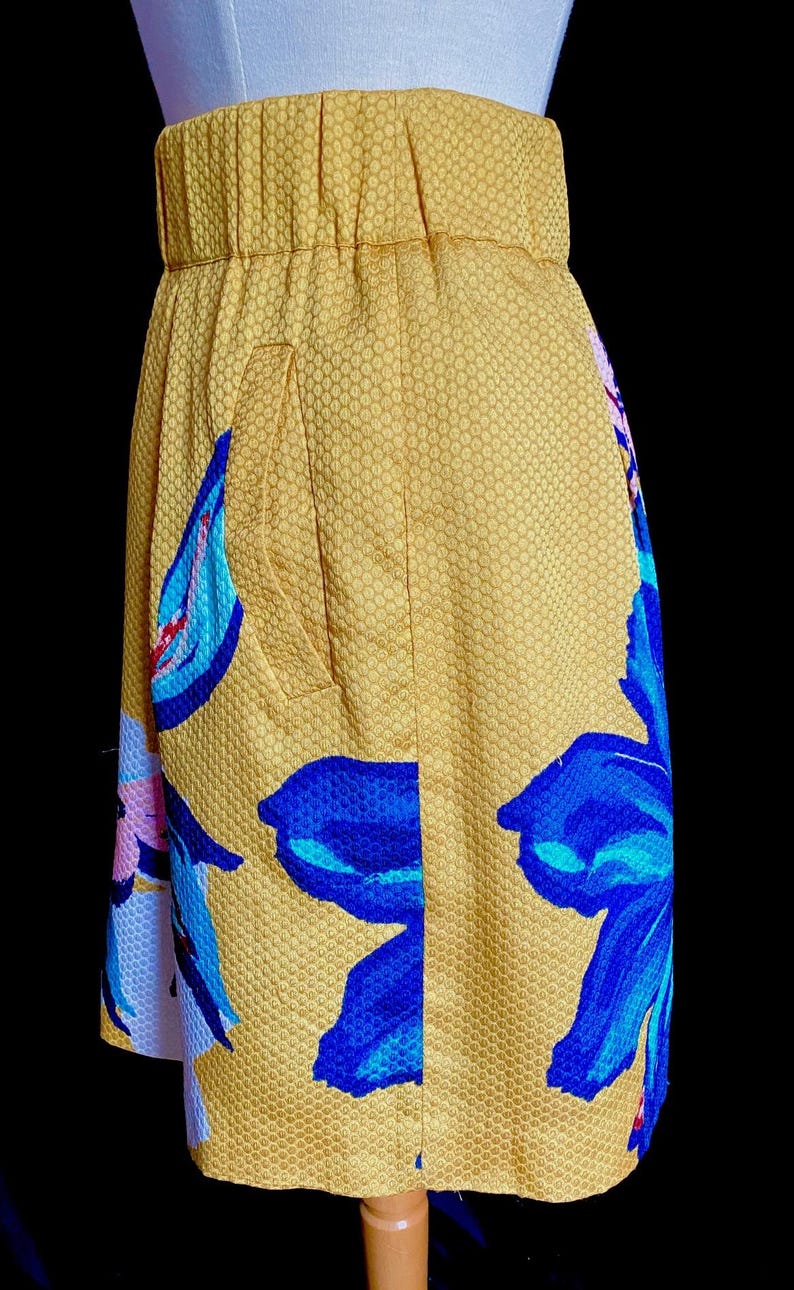 Op de afbeelding: Een gele rok met een bloemenpatroon. De rok heeft grote blauwe en roze bloemen op een gele, getextureerde achtergrond. De rok heeft een getailleerde tailleband en een zak.