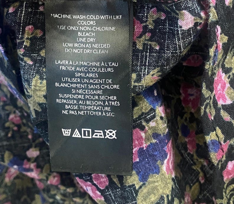 May include: Close-up of a black fabric with a floral pattern in pink, blue, and green. A black tag with white text details washing instructions: machine wash cold, non-chlorine bleach, line dry, low iron, and do not dry clean.