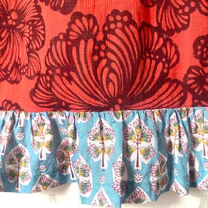 May include: Close-up of a fabric with a floral pattern. The top section is a vibrant red with large, dark burgundy floral designs. The bottom features a light blue ruffle with a repeating tree and floral pattern.