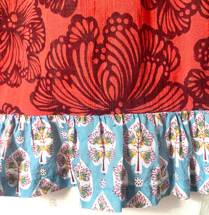 May include: Close-up of a fabric with a floral pattern. The top section is a vibrant red with large, dark burgundy floral designs. The bottom features a light blue ruffle with a repeating tree and floral pattern.