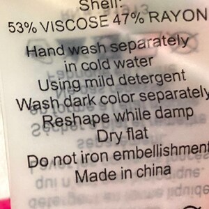 May include: A white clothing tag with black text. The tag includes care instructions: hand wash separately in cold water, reshape while damp, and dry flat. The tag also states the shell is 53% viscose and 47% rayon.