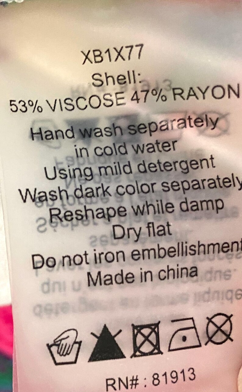 May include: A white clothing tag with black text. The tag includes care instructions: hand wash separately in cold water, reshape while damp, and dry flat. The tag also states the shell is 53% viscose and 47% rayon.