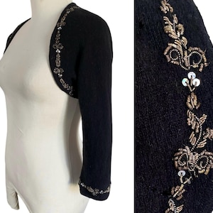 May include: Black bolero sweater with three-quarter sleeves. The sweater is embellished with intricate gold and silver beading and sequins along the edges and sleeves. The bolero is displayed on a mannequin.