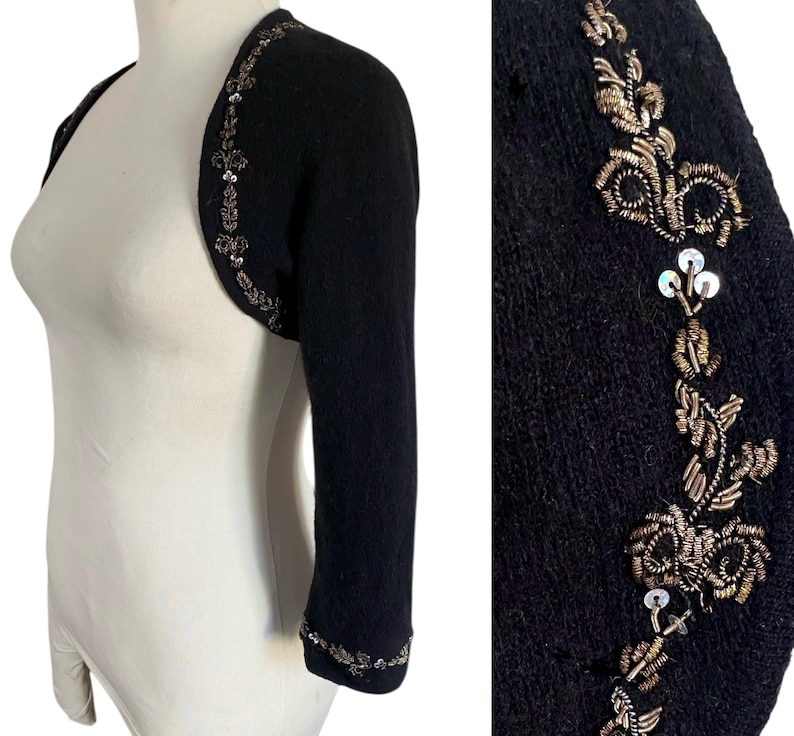 May include: Black bolero sweater with three-quarter sleeves. The sweater is embellished with intricate gold and silver beading and sequins along the edges and sleeves. The bolero is displayed on a mannequin.