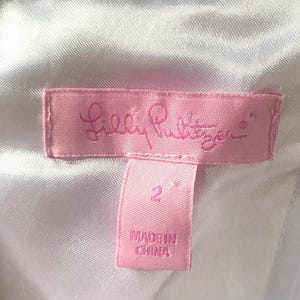 Op de afbeelding: Een roze label met het Lilly Pulitzer-logo in cursief schrift. Daaronder staat een kleiner roze label met het nummer "2" en de tekst "MADE IN CHINA". De achtergrond is een witte, getextureerde stof.