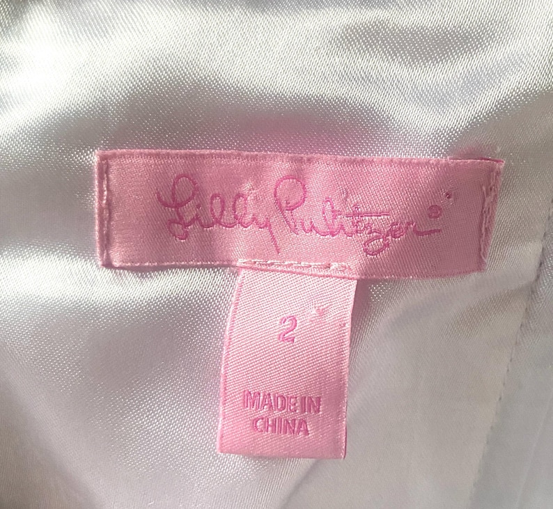Op de afbeelding: Een roze label met het Lilly Pulitzer-logo in cursief schrift. Daaronder staat een kleiner roze label met het nummer "2" en de tekst "MADE IN CHINA". De achtergrond is een witte, getextureerde stof.