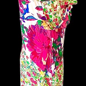 May include: A floral print dress on a mannequin. The dress features a white background with large pink flowers and smaller red, green, and blue floral accents. The dress has a ruffled neckline and a fitted silhouette.