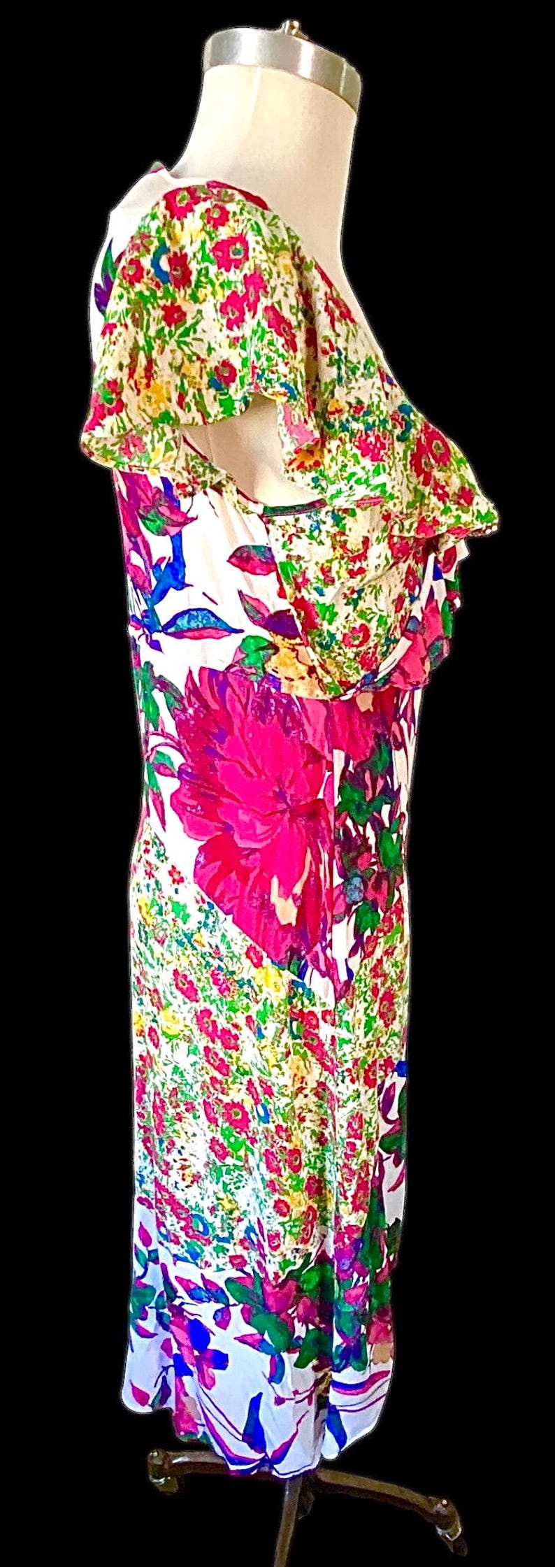 May include: A floral print dress on a mannequin. The dress features a white background with large pink flowers and smaller red, green, and blue floral accents. The dress has a ruffled neckline and a fitted silhouette.