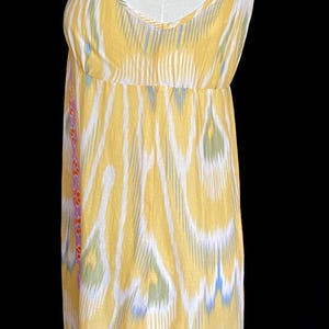May include: A sleeveless yellow dress with a white, blue, and green ikat pattern. The dress has a fitted bodice and a flowing skirt. The hemline and side seams are trimmed with a floral pattern in pink, orange, and yellow.