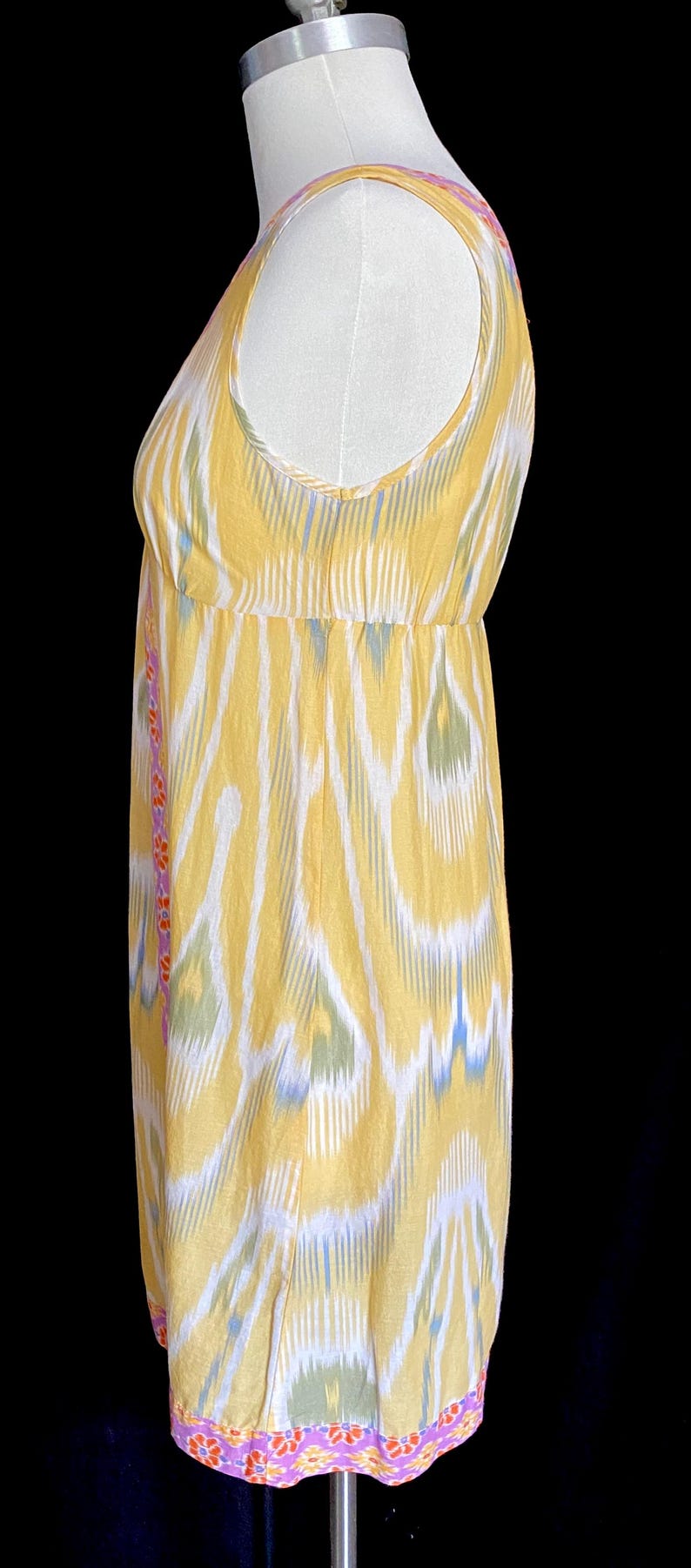 May include: A sleeveless yellow dress with a white, blue, and green ikat pattern. The dress has a fitted bodice and a flowing skirt. The hemline and side seams are trimmed with a floral pattern in pink, orange, and yellow.