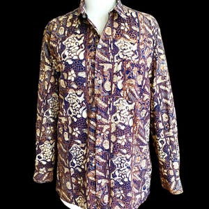 May include: A long-sleeved button-down shirt with a batik pattern. The shirt features a dark blue background with a repeating pattern of white, brown, and gray floral and geometric shapes. The shirt has a classic collar and buttoned cuffs.