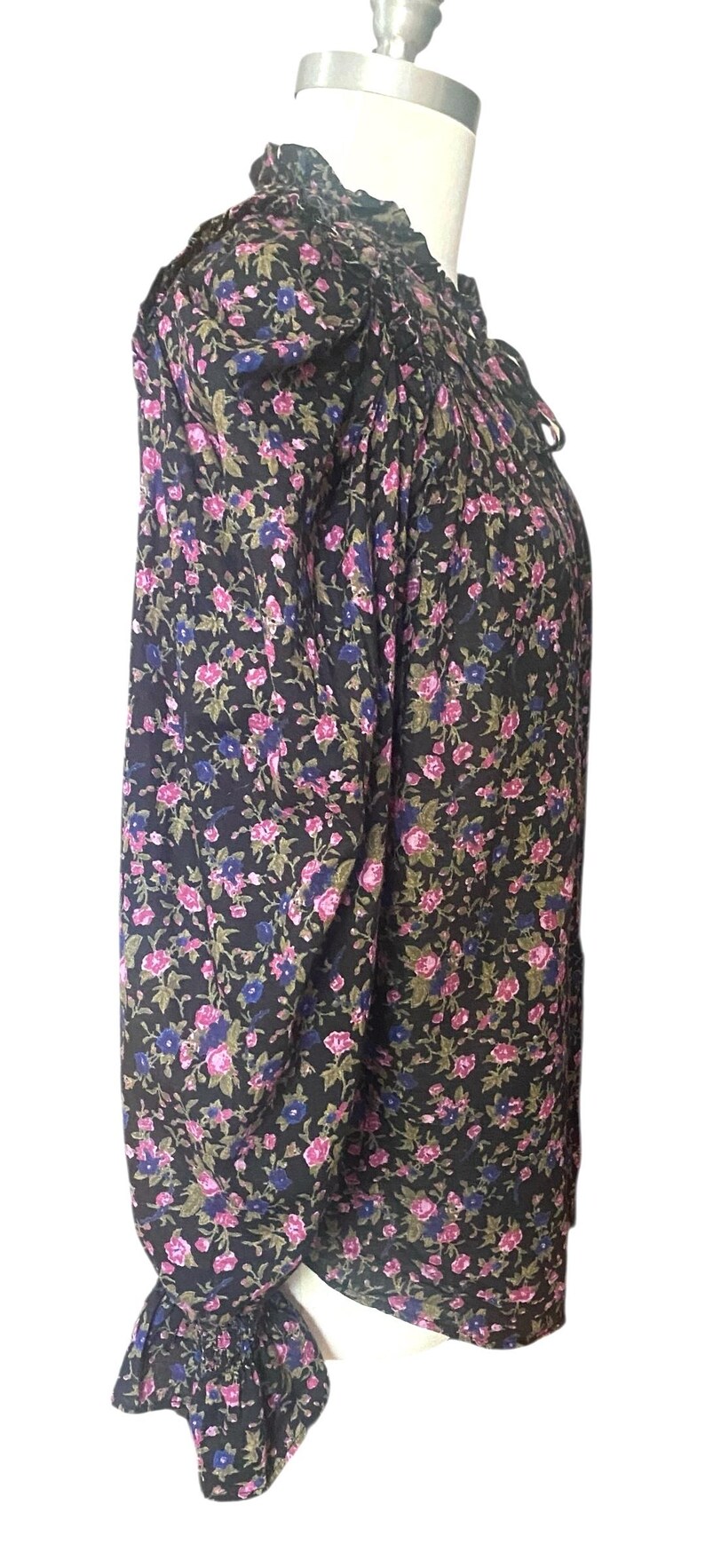 May include: A long-sleeved blouse with a floral pattern. The black fabric is covered in pink, blue, and green flowers. The blouse has a high ruffled collar and ruffled cuffs. The garment is displayed on a mannequin.