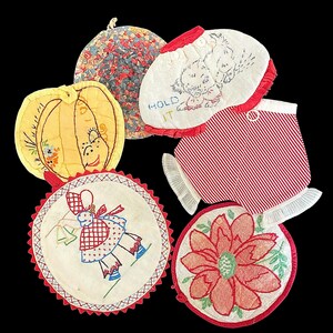 May include: A collection of six quilted fabric potholders featuring various designs including a yellow pumpkin, a floral pattern, a red and white striped piece, a circular embroidered figure, a red flower, and a white piece with the text HOLD IT.