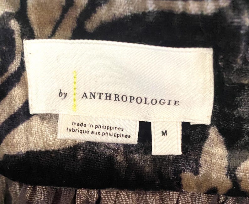 Puede incluir: Una etiqueta blanca con texto negro que dice "by ANTHROPOLOGIE" y "made in philippines/fabriqu&eacute; aux philippines". La etiqueta tambi&eacute;n tiene una etiqueta de talla que dice "M".