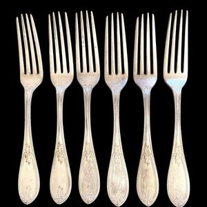 May include: A set of six silver-toned metal forks arranged in a row. Each fork features four tines and a handle decorated with an embossed floral scroll pattern at the base. The metal has a polished finish with visible signs of age and wear.