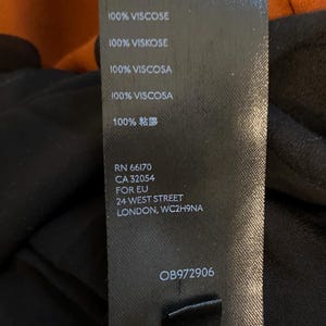 以下が含まれることがあります： 黒い衣類タグ。白い文字で「100% VISCOSE」が複数回繰り返されています。タグには登録番号、住所、製品コードも記載されています。背景には、オレンジと黒のドット柄の生地が写っています。