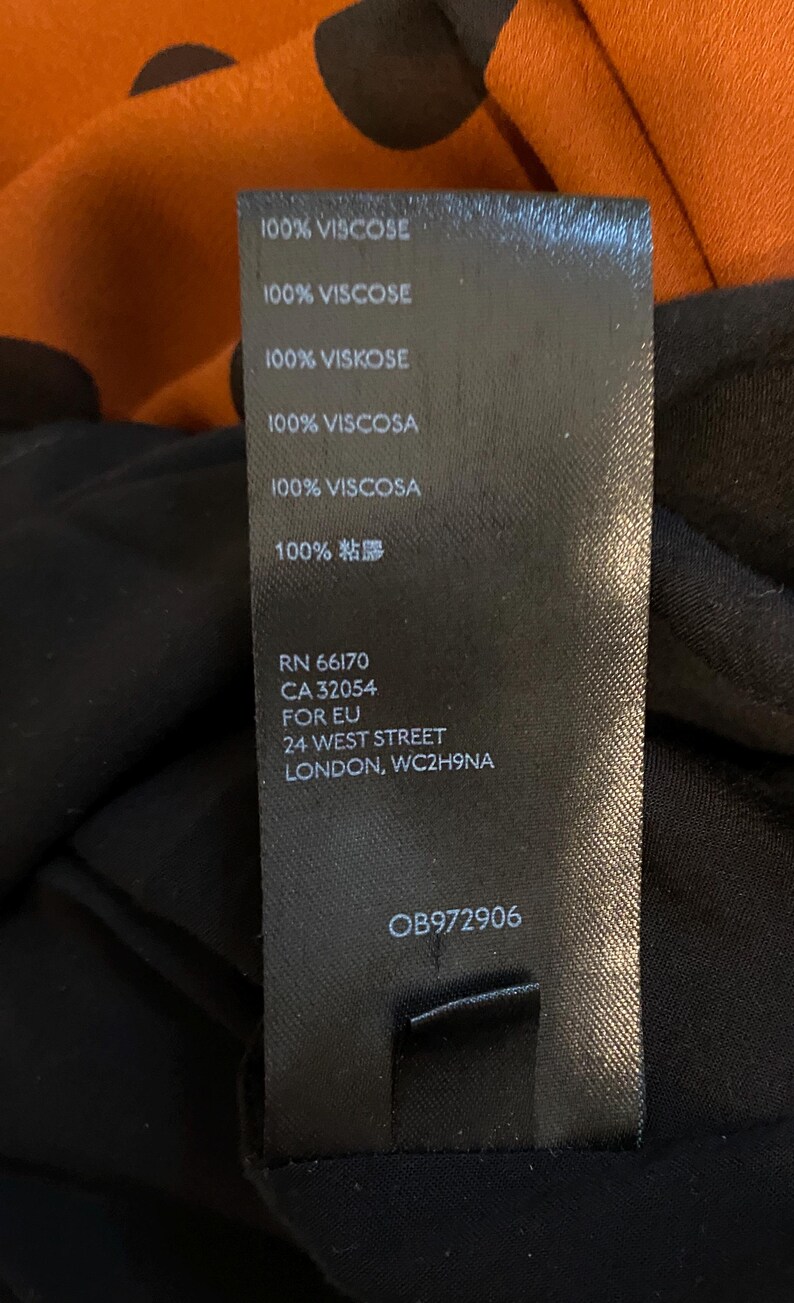 以下が含まれることがあります： 黒い衣類タグ。白い文字で「100% VISCOSE」が複数回繰り返されています。タグには登録番号、住所、製品コードも記載されています。背景には、オレンジと黒のドット柄の生地が写っています。