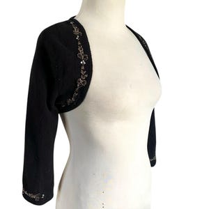 May include: Black bolero sweater with three-quarter sleeves. The sweater features intricate sequin and bead embellishments along the edges of the sleeves and neckline. The garment is displayed on a white mannequin.