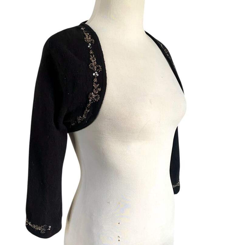 May include: Black bolero sweater with three-quarter sleeves. The sweater features intricate sequin and bead embellishments along the edges of the sleeves and neckline. The garment is displayed on a white mannequin.