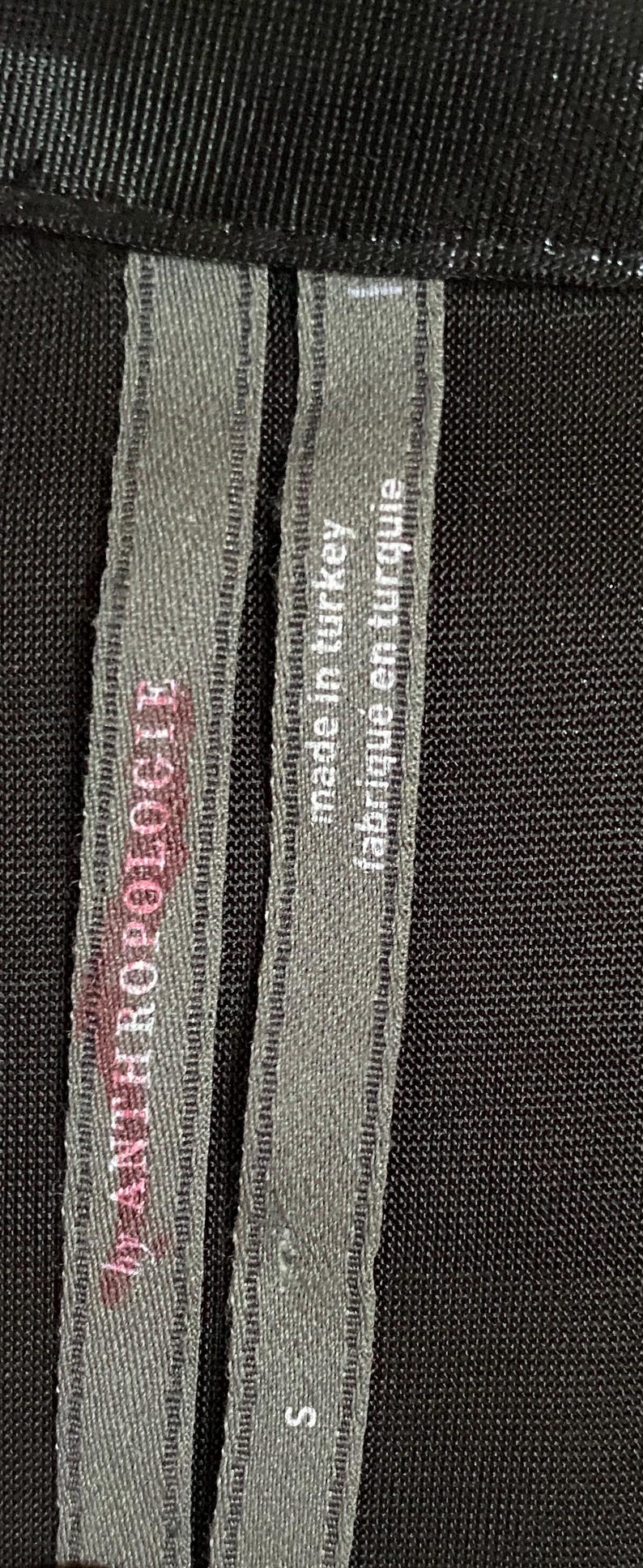 May include: A black fabric label with white text that reads "Made in Turkey, fabrique en turquie". The label also has the text "MANZERO ROGER" and "S" printed on it.