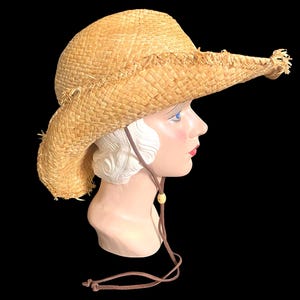 May include: A straw cowboy hat with a wide brim and frayed edges. The hat is light brown and has a brown leather chin strap with a wooden bead. The hat is displayed on a mannequin head against a black background.