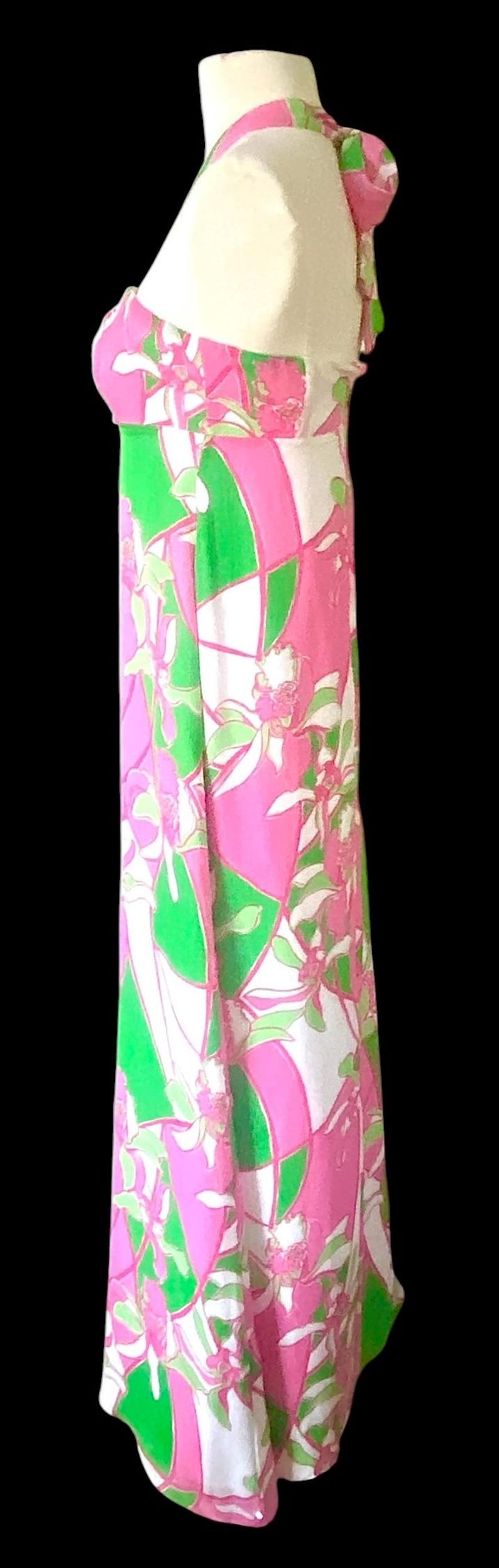 Op de afbeelding: Lange mouwloze jurk met een levendig bloemenpatroon in roze, groen en wit. De jurk heeft een halterhalslijn en een vloeiend silhouet. Het ontwerp bevat geometrische vormen en bloemmotieven.