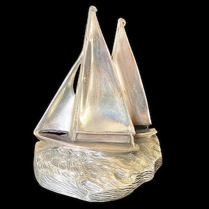 May include: A silver-colored sailboat sculpture with two sails. The boat sits atop a textured base, resembling ocean waves. The sculpture is made of a shiny metal and is set against a black background.