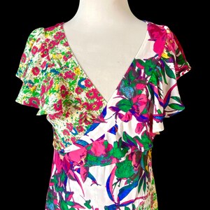 May include: A white V-neck dress with a vibrant floral print. The dress features a mix of pink, green, blue, and yellow flowers. The dress has ruffled sleeves and a fitted bodice.