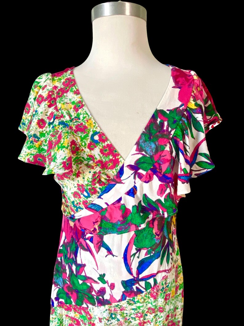 May include: A white V-neck dress with a vibrant floral print. The dress features a mix of pink, green, blue, and yellow flowers. The dress has ruffled sleeves and a fitted bodice.