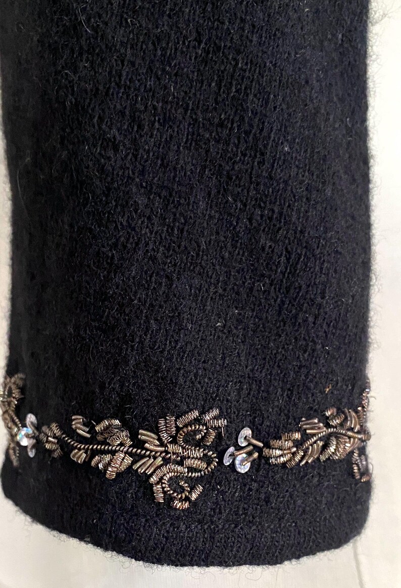 May include: Close-up of a black garment with a decorative trim. The trim features a floral design made of metallic wire and small, clear sequins. The fabric has a textured appearance.
