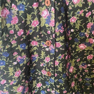 May include: Close-up of a dark green floral print fabric. The pattern features pink and blue flowers with green leaves. The fabric is buttoned down the front with brown buttons. The floral design is repeated throughout the fabric.
