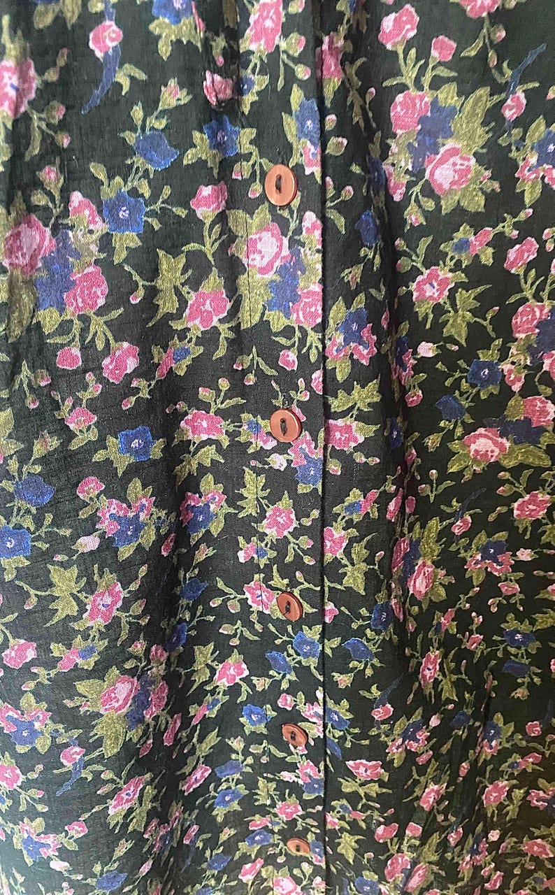 May include: Close-up of a dark green floral print fabric. The pattern features pink and blue flowers with green leaves. The fabric is buttoned down the front with brown buttons. The floral design is repeated throughout the fabric.
