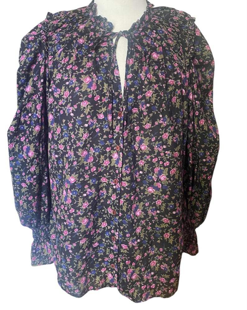 May include: A black floral print blouse with pink and blue flowers. The long-sleeved top has a ruffled collar and cuffs, and a button-down front. The blouse is made of a lightweight fabric.