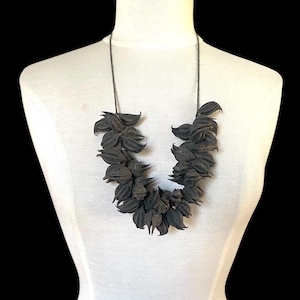 May include: A statement necklace featuring a cluster of dark, textured, pod-like organic elements arranged in a V-shape. The dark brown, pointed botanical components are strung together on a thin, dark cord, creating a dense, layered appearance.