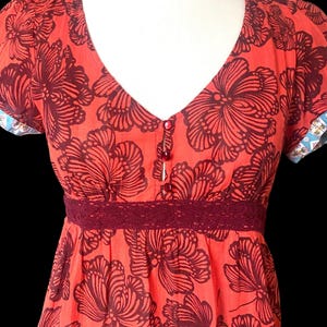 May include: A coral-colored dress with a V-neck and short sleeves. The dress features a large, dark burgundy floral pattern. A burgundy lace trim accents the waist. The sleeves have a patterned trim. The dress has three small buttons down the front.