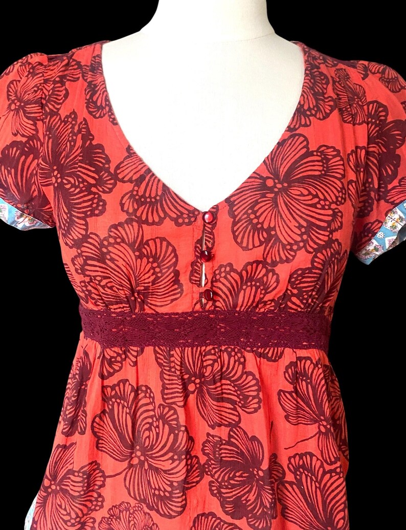 May include: A coral-colored dress with a V-neck and short sleeves. The dress features a large, dark burgundy floral pattern. A burgundy lace trim accents the waist. The sleeves have a patterned trim. The dress has three small buttons down the front.