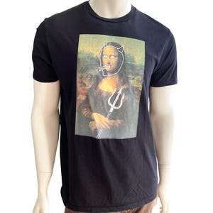 May include: Black t-shirt featuring a humorous graphic of the Mona Lisa with devil horns, a cigarette, and a trident. The graphic is a parody of the famous painting. The shirt is made of a soft material.